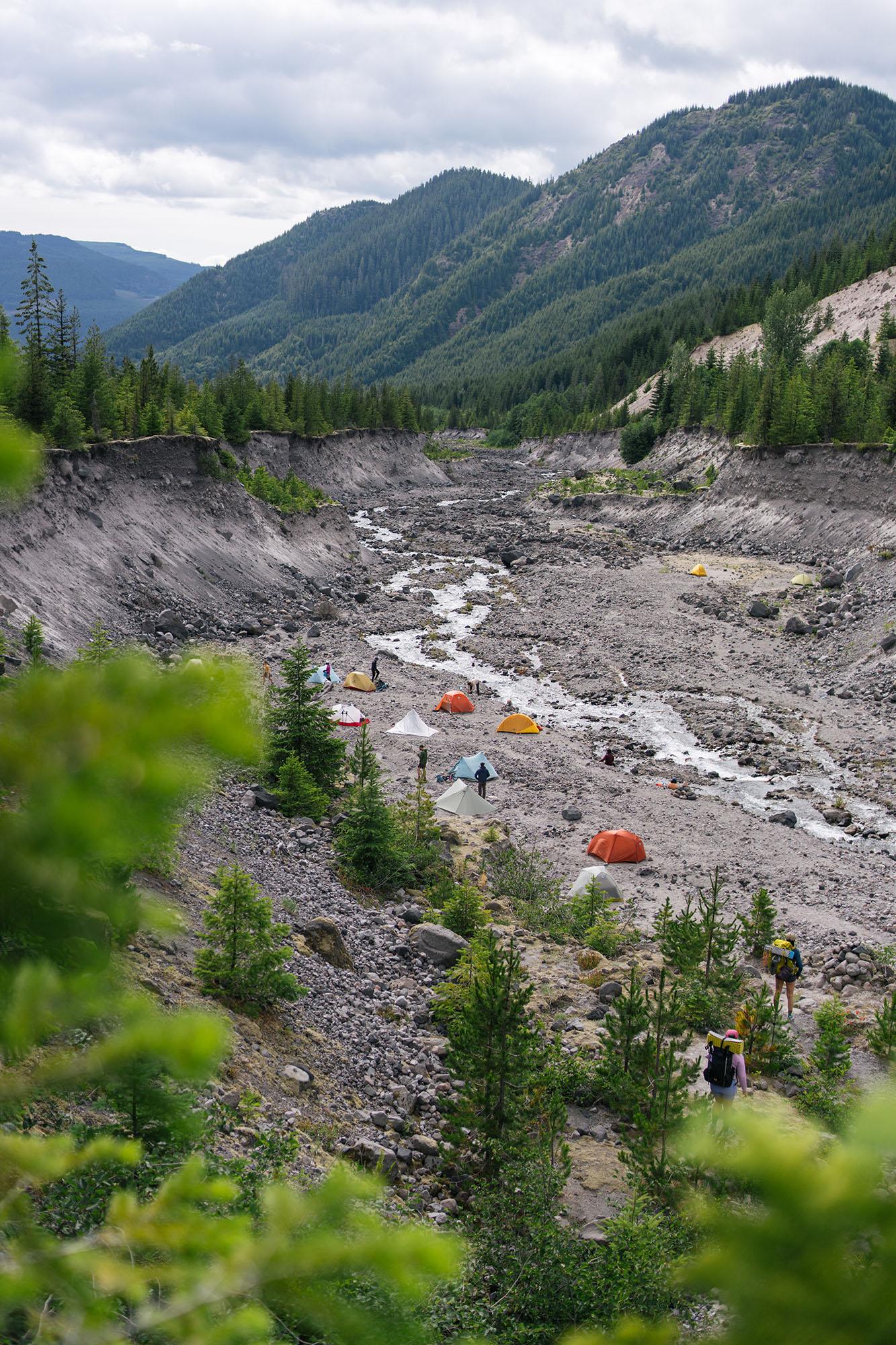 A large, rocky riverbed with a small creek running through it populated by a number of dome tents